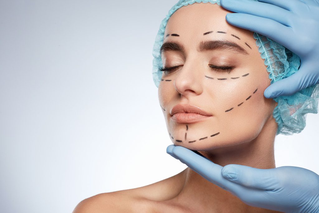 Beauty portrait of woman with closed eyes, plastic surgery concept, studio. Head and shoulders of model in blue cap with puncture lines on face, hands in blue gloves holding face